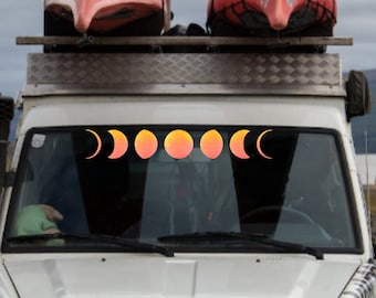 Puede incluir: Un todoterreno blanco con baca que transporta dos canoas rojas. El parabrisas muestra una calcomanía de las fases lunares en tonos naranja y amarillo. El vehículo está estacionado cerca de un lago.