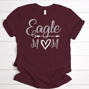 Puede incluir: Camiseta color burdeos con un gr&aacute;fico plateado que dice "Eagle Mom" con una flecha y un coraz&oacute;n.