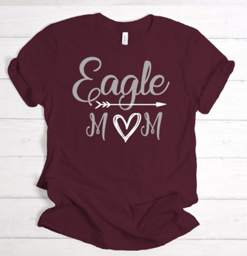 Puede incluir: Camiseta color burdeos con un gr&aacute;fico plateado que dice "Eagle Mom" con una flecha y un coraz&oacute;n.