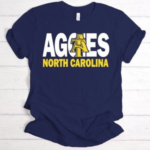 May include: Navy blue t-shirt with white text that reads "Aggies North Carolina". The "A" in Aggies is a yellow and gold stylised letter.