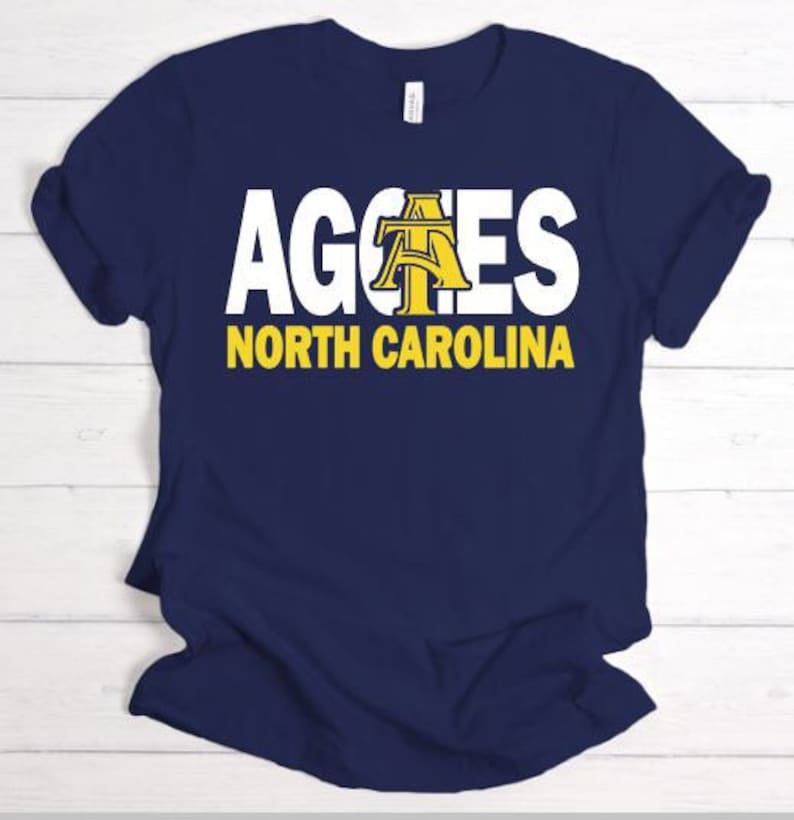 May include: Navy blue t-shirt with white text that reads "Aggies North Carolina". The "A" in Aggies is a yellow and gold stylised letter.