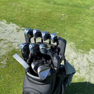 May include: A black golf bag containing a set of silver metal golf clubs. The clubs are arranged in the bag with the iron heads facing upward, including two wedges clearly labeled with the text HI-TOE and 54, and 58, respectively.