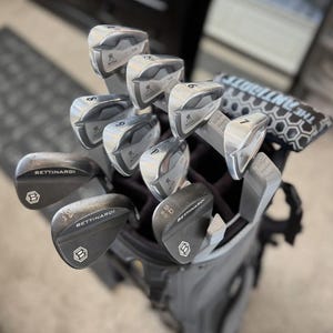 May include: A set of golf clubs in a gray golf bag. The clubs include silver irons with the "Miura" logo and black wedges with the "Bettinardi" logo. The bag also contains a black and white head cover.