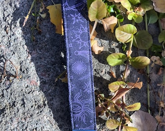 May include: A dark blue key fob with a celestial pattern of planets, stars, and suns in purple. The key fob has a silver metal clasp and ring. The background is a textured gray surface with green and brown foliage.