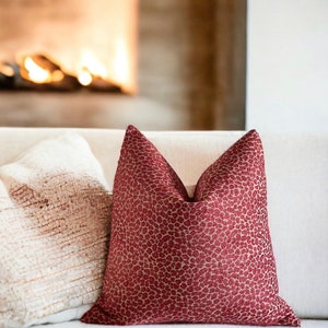 May include: A red and beige patterned throw pillow with a subtle leopard print design. The pillow is sitting on a white couch in front of a fireplace.