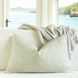 May include: A decorative throw pillow with a white background and a pattern of small, light green dots. A light gray throw blanket with fringe is draped over the back of the sofa. The sofa is white, and the background shows a view of the ocean.