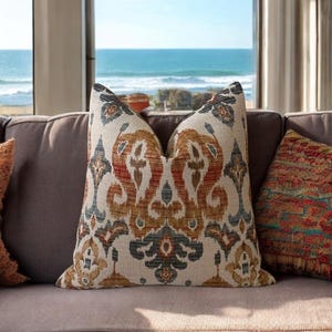 May include: A brown couch with two throw pillows. The couch is in front of a window with a view of the ocean. The throw pillows have a colorful geometric pattern.
