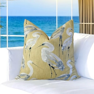 May include: A decorative throw pillow featuring a repeating pattern of white and gray herons on a golden-yellow background. The pillow is placed on a white surface, with a view of the ocean in the background.