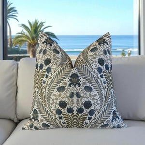 May include: A square throw pillow featuring a symmetrical botanical pattern with dark blue and olive green floral motifs and leafy stems on a white background. The fabric has a dense, intricate print of stylized flowers and foliage.