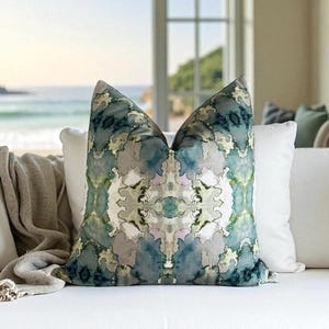 May include: A decorative throw pillow with a symmetrical pattern in shades of blue, green, gray, and white. The pillow is on a white sofa with a beach scene visible in the background. The pillow has a soft, textured appearance.