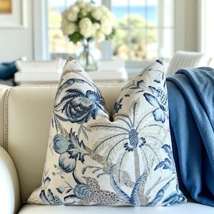 May include: A decorative throw pillow with a botanical design in shades of blue, gray, and cream. The pillow features a detailed pattern of flowers, foliage, and a palm tree. The pillow is placed on a white sofa next to a blue blanket.