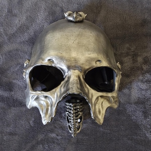 Puede incluir: Máscara de calavera plateada con grandes aberturas para los ojos y una mandíbula esquelética. La máscara tiene una superficie texturizada y un pequeño elemento decorativo en la parte superior. La máscara es probablemente para disfraces o decoración.