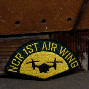 May include: A green and yellow embroidered patch with the text "NCR 1ST AIR WING" in yellow lettering. A black silhouette of a helicopter is in the centre of the patch.