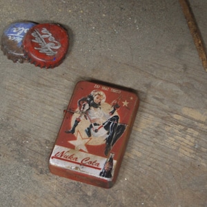 May include: A red and silver Nuka Cola lighter with a pin-up girl graphic and the text "Zap That Thirst!" and "Nuka Cola". Two bottle caps are also visible, one red and one blue.