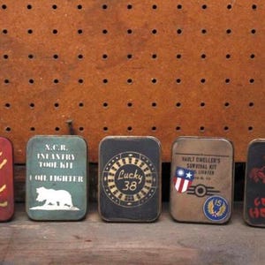 May include: Five rectangular metal tins with diverse designs and text. The tins are in shades of red, green, black, and brown. The designs include a yellow silhouette, a white bear, a roulette wheel, an American flag, and a red horse. The text includes "N.C.R. Infantry Tool Kit", "Lucky 38", and "Vault Dweller's Survival Kit".