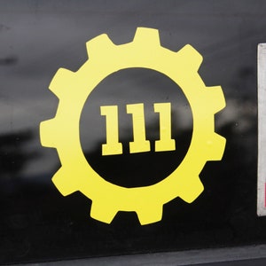 May include: A yellow gear-shaped sticker with the number "111" inside. The sticker is on a dark surface, with a portion of a California state flag visible to the right. The gear has a cogwheel design.