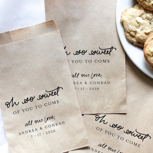 Può includere: Sacchetti regalo di carta marrone con la scritta "Oh so sweet, of you to come, all our love, Andrea & Conrad, 7.17.2026" in corsivo nero. A destra un piatto di biscotti con gocce di cioccolato bianco.