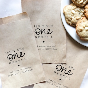May include: Brown paper party favor bags with the text "ISN'T SHE ONE DERFUL" and "thank you for coming! OLIVIA'S 1st BIRTHDAY" printed in black. A plate of white chocolate chip cookies is visible. The bags are for a first birthday celebration.