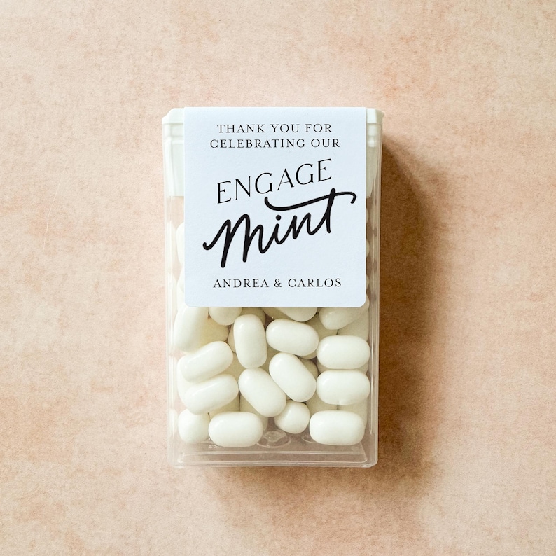 May include: A clear plastic box filled with white mints. The box has a white label with the text "THANK YOU FOR CELEBRATING OUR ENGAGE MINT ANDREA & CARLOS" in black script. The mints are oval-shaped.