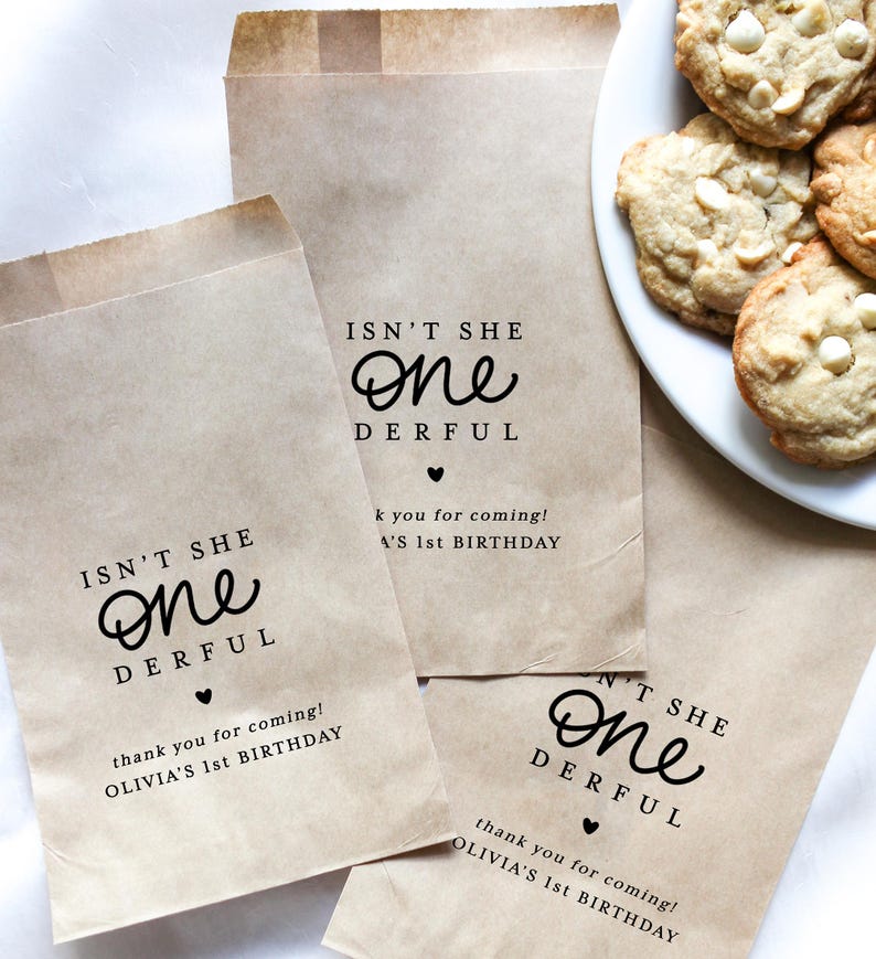 May include: Brown paper party favor bags with the text "ISN'T SHE ONE DERFUL" and "thank you for coming! OLIVIA'S 1st BIRTHDAY" printed in black. A plate of white chocolate chip cookies is visible. The bags are for a first birthday celebration.