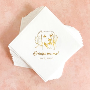 May include: A stack of square white paper napkins featuring a gold foil illustration of a dog's face. Below the dog, the text reads "Drinks on me!" in a cursive script, followed by the text "LOVE, ARLO" in a simple, clean sans-serif font.