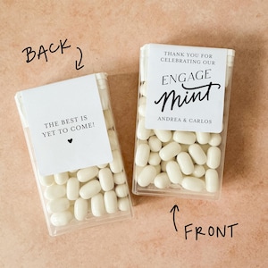 May include: Two clear plastic boxes filled with white mints. One box reads "THE BEST IS YET TO COME!" and the other says "ENGAGE mint" with names. The boxes are on a light brown surface.