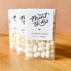 May include: Three clear plastic containers filled with white mints. Each container has a white label with the text "Mint to Be", a heart, and the names "Alexa & James" with the date "6.3.2026". The containers are on a wooden surface.
