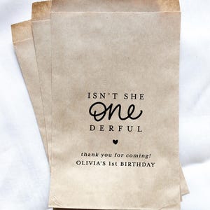 May include: Three kraft paper favor bags with the text "Isn't She One Derful" and "thank you for coming! OLIVIA'S 1st BIRTHDAY" printed in black. The bags are stacked on top of each other.