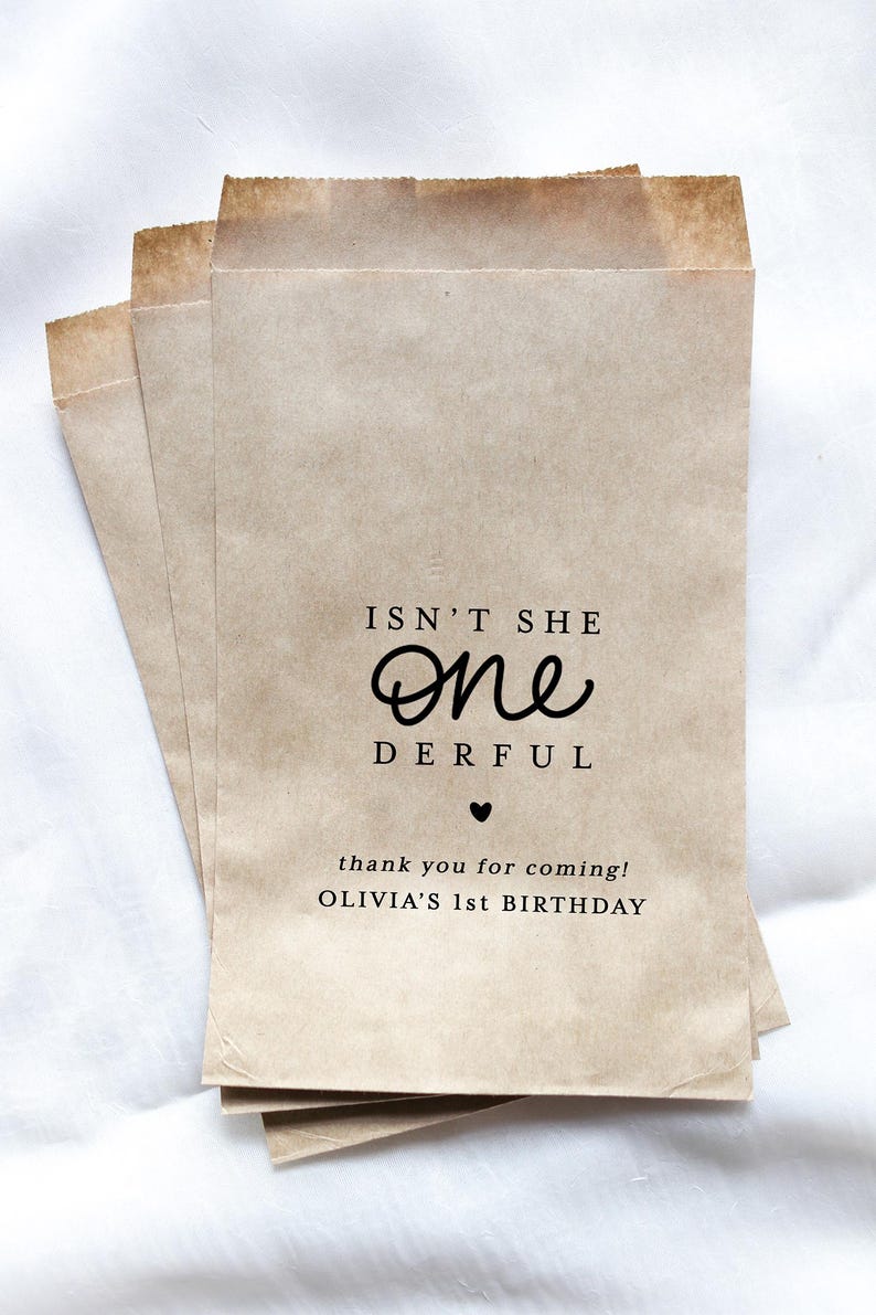 May include: Three kraft paper favor bags with the text "Isn't She One Derful" and "thank you for coming! OLIVIA'S 1st BIRTHDAY" printed in black. The bags are stacked on top of each other.