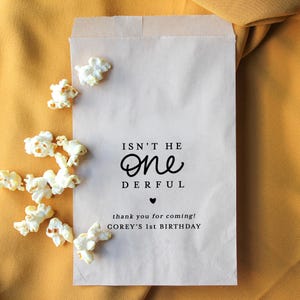 May include: A light brown paper bag with the text "ISN'T HE ONE DERFUL" and "thank you for coming! COREY'S 1st BIRTHDAY". Scattered popcorn kernels are visible on a yellow background.
