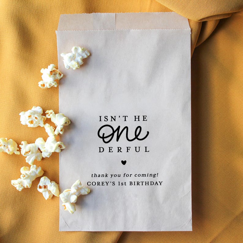 May include: A light brown paper bag with the text "ISN'T HE ONE DERFUL" and "thank you for coming! COREY'S 1st BIRTHDAY". Scattered popcorn kernels are visible on a yellow background.