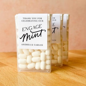May include: Clear plastic boxes filled with white mints. Each box has a white label with the words "THANK YOU FOR CELEBRATING OUR ENGAGE MINT" and the names "ANDREA & CARLOS" in black script. The boxes are arranged on a wooden surface.