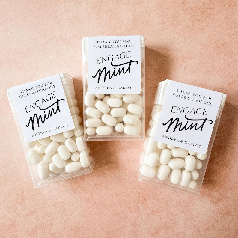 May include: Three transparent boxes filled with white mints. Each box has a white label with the words "THANK YOU FOR CELEBRATING OUR ENGAGE Mint" and the names "ANDREA & CARLOS" in black script.