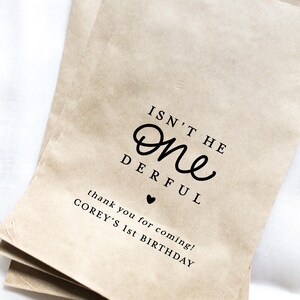 May include: Three brown paper party favor bags with the text "ISN'T HE One DERFUL" and "thank you for coming! COREY'S 1st BIRTHDAY" printed in black. A small black heart is between the phrases.