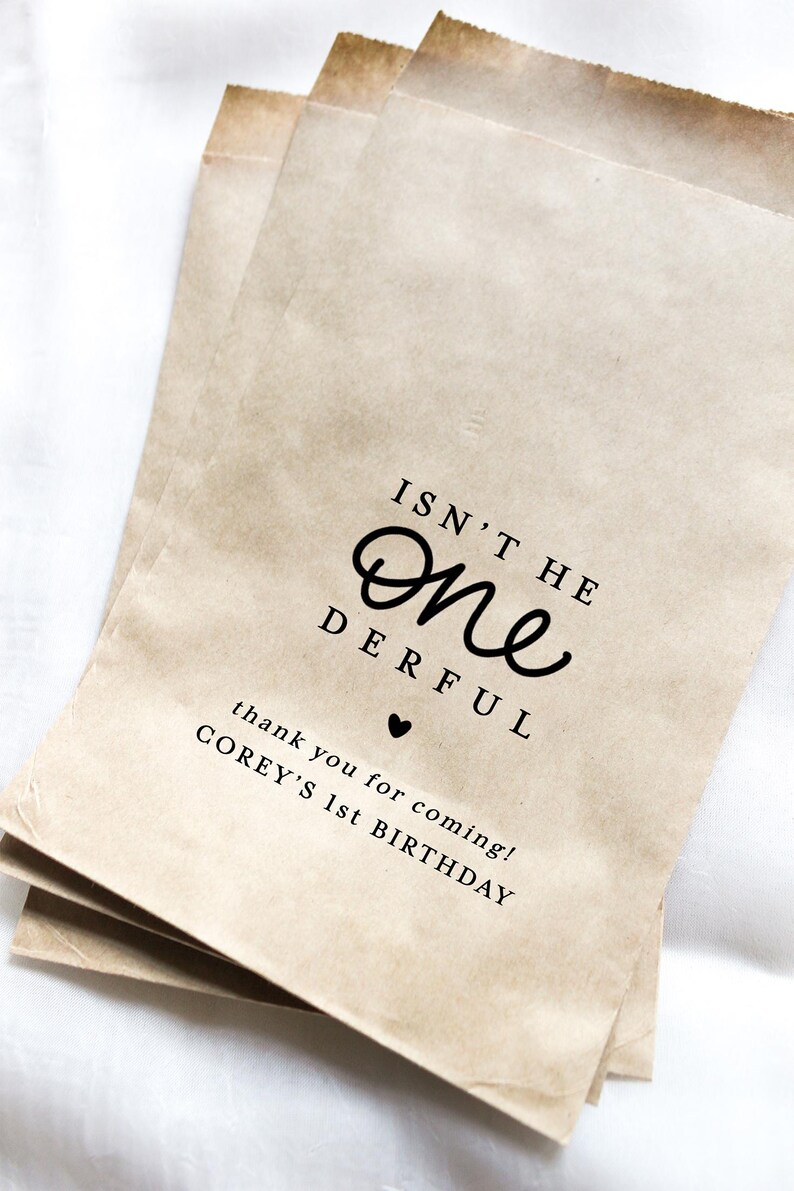 May include: Three brown paper party favor bags with the text "ISN'T HE One DERFUL" and "thank you for coming! COREY'S 1st BIRTHDAY" printed in black. A small black heart is between the phrases.