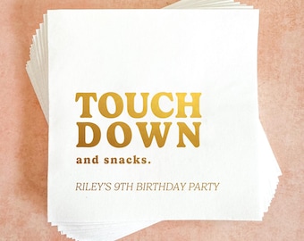 May include: A stack of square white paper napkins featuring gold foil text that reads TOUCH DOWN and snacks. below which is the text RILEY'S 9TH BIRTHDAY PARTY in a smaller font. The napkins are arranged in a slightly offset, layered pile.