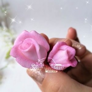 Puede incluir: Dos moldes de jabón con forma de rosa rosa. Los moldes se sostienen en una mano. Los moldes están hechos de silicona y están diseñados para crear rosas de aspecto realista.
