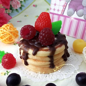 Op de afbeelding: Een stapel mini-pannenkoeken bedekt met chocoladesaus en rode frambozen. De pannenkoeken liggen op een wit servet met blauwe bessen en frambozen die eromheen zijn verspreid.