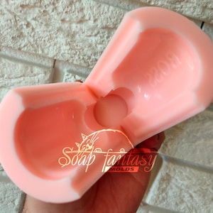 May include: A pink silicone mould for making soap. The mould is shaped like a heart with a round hole in the centre. The mould is open and the inside is visible. The mould has the text "Soap Fantasy Molds" printed on the bottom.