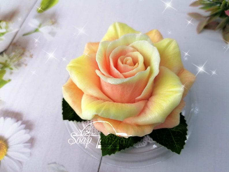 May include: A handcrafted soap shaped like a rose. The soap features a gradient of colors, with a peach center that fades to yellow on the outer petals. Green leaves and the text "Soap Te" are visible.