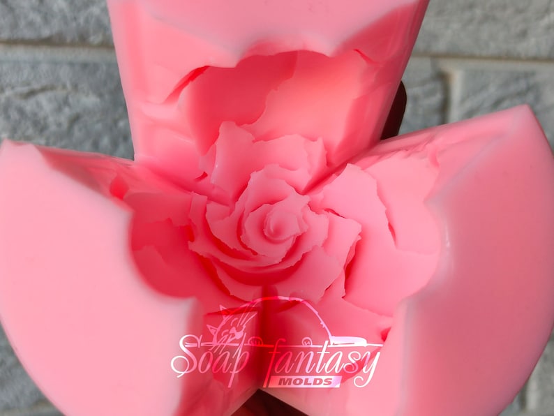 May include: Pink silicone mold shaped like a rose. The mold is open and shows the detailed rose design. The mold is labeled "Soap Fantasy Molds".