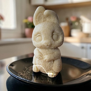 May include: A cream-colored bunny figurine with large, open eyes and folded arms, sitting on a black, oval-shaped plate. The bunny has textured fur and large ears. The plate has a decorative rim and is placed on a dark surface. The background is blurred.