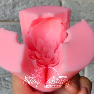 May include: Pink silicone mold for making a flower-shaped candle. The mold is open to show the detailed design of the flower petals. The mold is labeled "Soap Fantasy Molds".