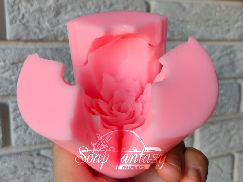 May include: Pink silicone mold for making a flower-shaped candle. The mold is open to show the detailed design of the flower petals. The mold is labeled "Soap Fantasy Molds".