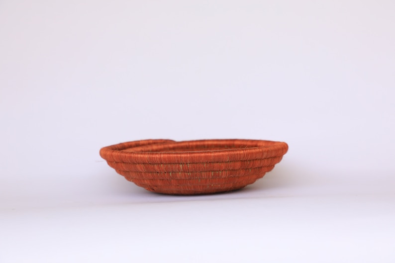 Pode incluir: Uma cesta tecida de fibras naturais em uma cor laranja intensa. A cesta &eacute; redonda e tem uma profundidade rasa. &Eacute; perfeita para guardar objetos pequenos como joias ou bugigangas.