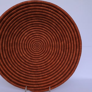 Pode incluir: Uma cesta grande, redonda e tecida com um padr&atilde;o em espiral. A cesta &eacute; feita de fibras naturais e &eacute; de cor castanho-avermelhado escuro.