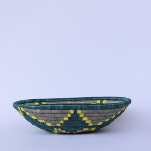 K&ouml;nnte beinhalten: Ein geflochtener Korb mit einem teal- und grauen Muster und gelben Akzenten. Der Korb ist oval geformt und hat eine geringe Tiefe.