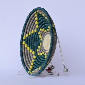 K&ouml;nnte beinhalten: Ein geflochtener Korb mit einem teal-, grau- und gelben Muster. Der Korb steht auf einem klaren Acryl-St&auml;nder.
