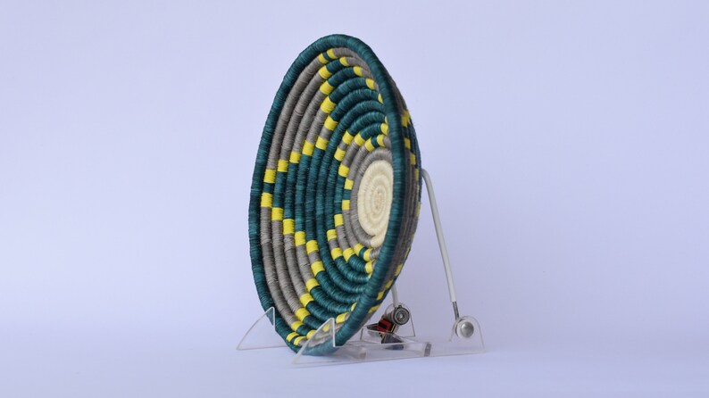 K&ouml;nnte beinhalten: Ein geflochtener Korb mit einem teal-, grau- und gelben Muster. Der Korb steht auf einem klaren Acryl-St&auml;nder.
