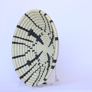 May include: A large, round woven basket with a black and white striped pattern. The basket is made of natural fibers and has a slightly raised rim. It is displayed on a clear acrylic stand.